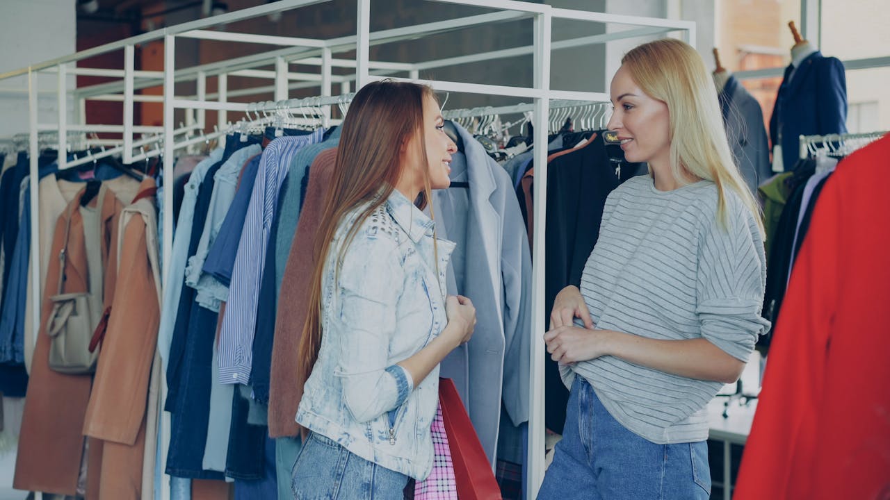 Two women chatting among clothing racks in a stylish fashion store, sharing shopping advice.